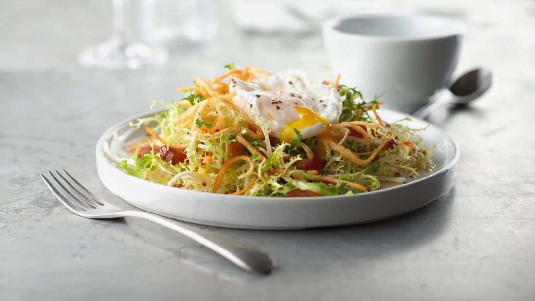 Salad Lyonnaise topped with a poached pasteurized shell egg, crispy hash browns, and bacon—premium egg add-on for foodservice menus